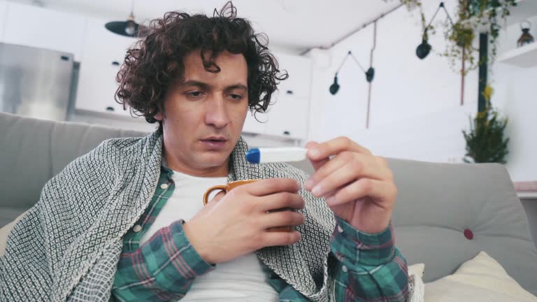 A sick man wrapped in a blanket on a sofa, holding a hot beverage and looking at a fever thermometer.