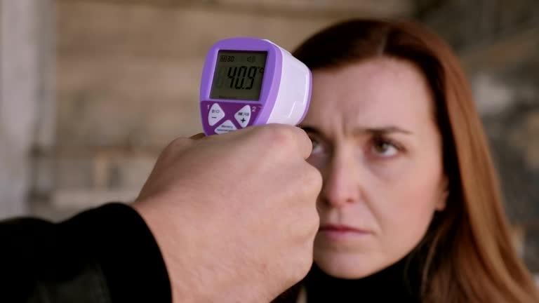 A hand holding a purple infrared non-contact fever thermometer to check a woman's forehead temperature, showing a high reading of 40.9 degrees.