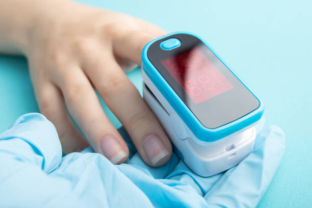 Close-up of a blue and white fingertip pulse oximeter placed on a person's finger, resting on a blue medical glove with a bright blue background.