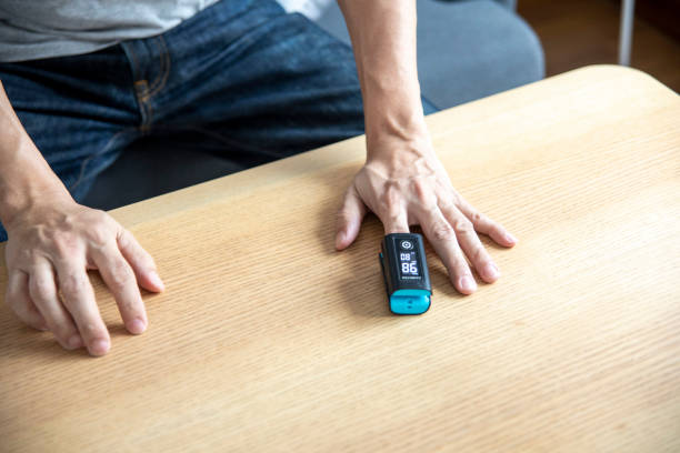 Top view of a man sitting at a wooden table, wearing a blue and black pulse oximeter on his index finger to measure oxygen levels.